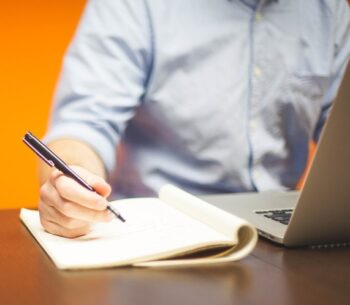 A person writing on paper with a laptop in the background.